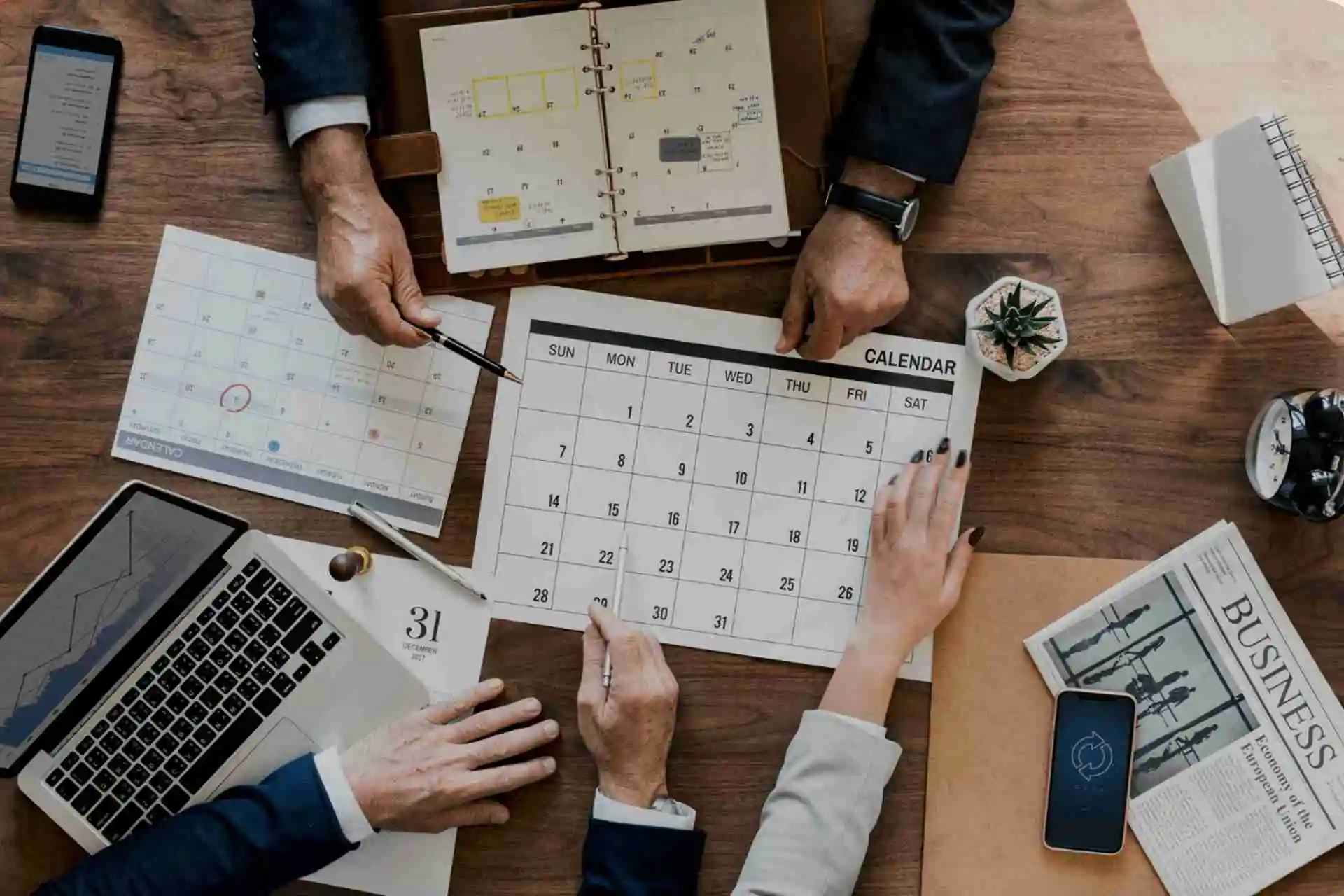 Profissionais de contabilidade planeando o cronograma mensal com um calendário numa mesa de reunião, rodeados por um portátil, relatórios financeiros e um jornal de negócios
